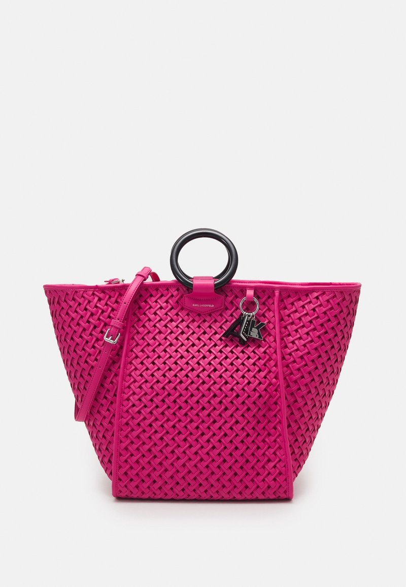 Sac à main tissé rose avec une large base, une poignée ronde noire, une sangle détachable et un porte-clés décoratif ; présente un matériau texturé et une forme structurée.