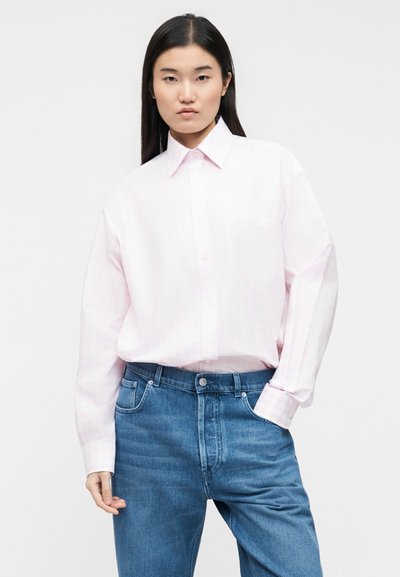 Νεαρή γυναίκα με μακριά σκούρα μαλλιά, φορώντας ένα oversized πουκάμισο σε απαλό ροζ χρώμα και μπλε τζιν, μπροστά σε απλό φόντο.
