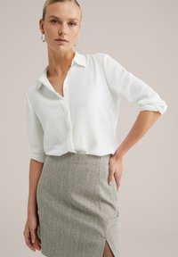 Una donna con i capelli raccolti indossa una camicia bianca con bottoni e maniche arrotolate, insieme a una gonna grigia a spina di pesce con uno spacco frontale, con la mano sulla vita.