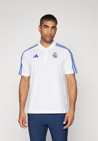 Polo bianco con strisce blu sulle maniche, con logo ricamato del Real Madrid e marchio Adidas. Il tessuto sembra liscio e leggero.