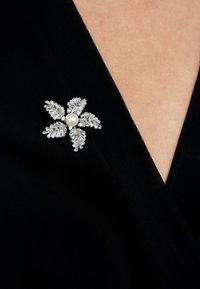 Silver flower brooch adorned with a central pearl, featuring leaf-like detailing and geometric accents, set against a black fabric background.