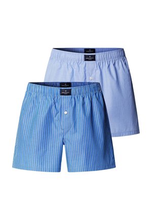 Joven hombre musculoso de pie descalzo, vestido con bóxers a rayas azules y blancas sobre un fondo gris claro liso.
