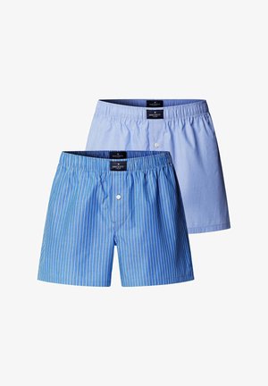 Joven hombre musculoso de pie descalzo, vestido con bóxers a rayas azules y blancas sobre un fondo gris claro liso.