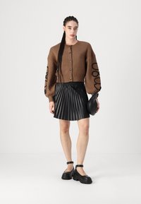 Barna kötött kardigán gombokkal, oversize ujjaival, fekete mintával; redőzött fekete szoknya; vastag talpú fekete cipők; fekete clutch táska.
