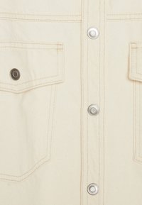 Beige denimskjorta med två bröstfickor, silverfärgade tryckknappar och kontrasterande sömmar som framhäver en texturerad tygdesign.