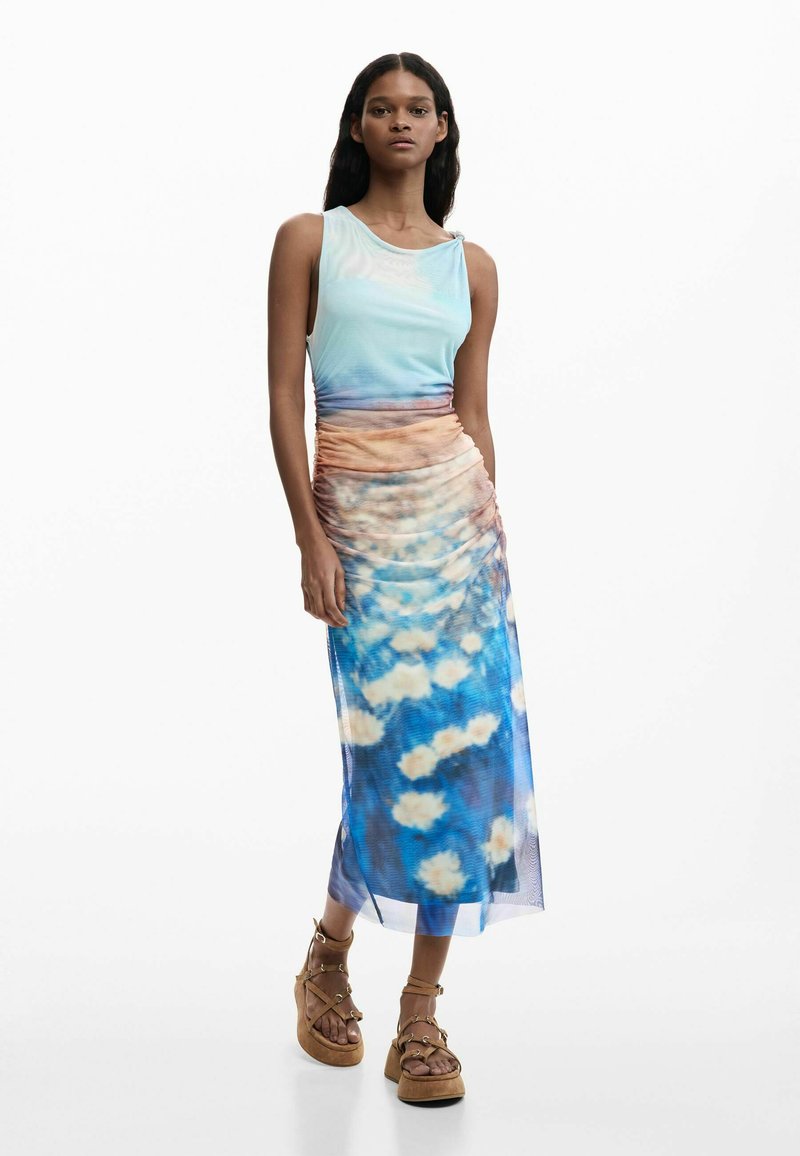 Frau steht und trägt ein ärmelloses Ombré-Kleid mit geraffter Taille und blauer Blumenkante, kombiniert mit beigefarbenen Riemchen-Plateau-Sandalen.