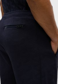 Pantaloni blu navy con motivo geometrico testurizzato, dotati di tasca posteriore con zip e tasche laterali. Tessuto morbido con taglio aderente.