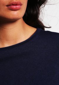 Marinblå stickad tröja med rundad halsringning och mjuk textur, delvis synlig med en bildvinkel från midjan uppåt, som framhäver slät hud.