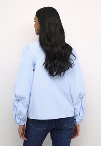 Camicia azzurro chiaro con strisce verticali, maniche a sbuffo e dettaglio arricciato ai polsini. Abbinata a jeans in denim scuri.