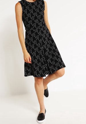 Mujer con vestido negro sin mangas hasta la rodilla con patrón geométrico blanco y zapatos negros sin cordones, de pie sobre una pierna.
