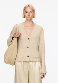 Beige Strickcardigan mit V-Ausschnitt, zwei Fronttaschen und Holzknöpfen. Kombiniert mit glänzenden, hellen Hosen und einer weichen beigefarbenen Tasche.