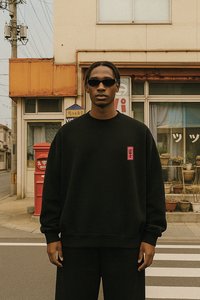 Νέος άνδρας με μαύρο φούτερ και γυαλιά ηλίου στέκεται σε διάβαση πεζών μπροστά από ένα παλαιό κτίριο με γλάστρες και κόκκινο ταχυδρομικό κουτί.