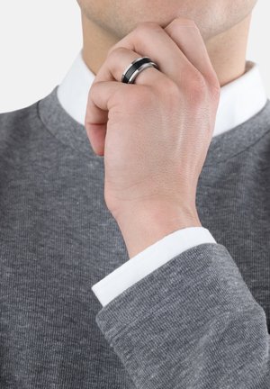 Black and silver ring worn on a hand, with a close-up of textured grey sweater and white collar. Focus on ring's design and finish.
