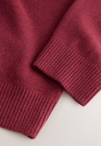 Detalje af en bordeaux uldtrøje med glat stoftstruktur og ribbet kant, der fremhæver en dyb farve og et blødt udseende.