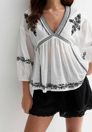 Witte blouse met zwarte bloemrijke borduursel, V-hals en driekwart mouwen. Gecombineerd met zwarte korte broek met een gegolfde zoom.