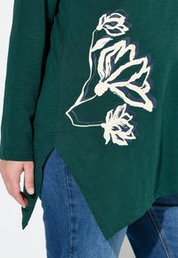 Grüner Langarmshirt mit einem weißen Blumenmuster auf der Vorderseite, mit abgerundetem Saum und seitlichen Schlitzen, kombiniert mit blauen Jeans.