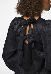 Blusa nera con maniche a sbuffo, realizzata in tessuto testurizzato, con scollatura a barco e due nastri di seta decorativi legati al colletto.