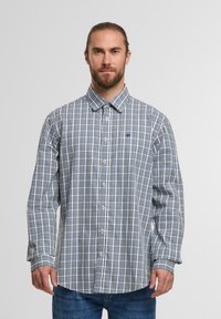 Mann mit Bart, der ein langärmliges, knöpfen besetztes, blau-weiß kariertes Hemd und eine blaue Jeans trägt, steht vor einem schlichten hellen Hintergrund.