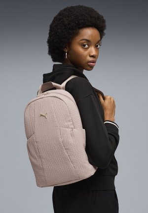 Jonge vrouw met een zwarte hoodie en een lichtroze, getextureerde Puma-rugzak over haar schouder tegen een grijze achtergrond.