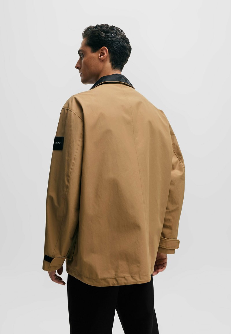 Beige oversized Jacke mit schwarzem Kragen, sichtbaren Nähten und einem Logo-Patch am Ärmel. Glatte Textur und lockere Passform.