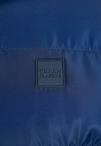 Mørkeblå, lett stoff med glatt tekstur og glans; har en firkantet svart lærpute preget med "URBAN CLASSICS."