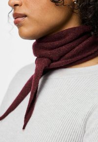 Burgundi spalvos šalikas, pagamintas iš minkštos medžiagos, surištas aplink kaklą. Fone matomas šviesiai pilkos spalvos pūkuotas megztinis.
