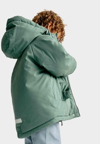 Zelená péřová bunda s velkou kapucí, zipem na přední straně a bočním kapsou. Vyrobena z měkkého, texturovaného materiálu s mírně lesklým povrchem.