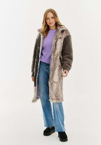 Žena v dlouhém, strukturovaném hnědo-krémovém kabátu přes fialový svetr, modré džíny a černé platformové boty, stojící na bílém pozadí.