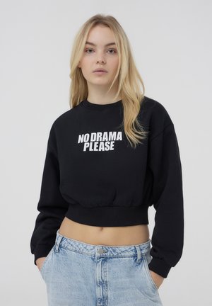 Felpa nera corta con testo bianco "NO DRAMA PLEASE". Presenta un collo rotondo e polsini a coste, abbinata a jeans azzurri effetto lavaggio acido.