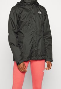 Veste noire imperméable avec capuche, dotée d'une fermeture éclair à l'avant et de poches. Le tissu est lisse avec une finition mate et le logo est présent sur la poitrine.