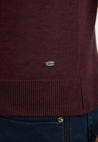 Bordeauxroter Strickpullover mit geripptem Saum, versehen mit einem kleinen silbernen Etikett mit der Aufschrift "Petrol" am unteren Rand. Getragen über dunkelblauen Jeans.