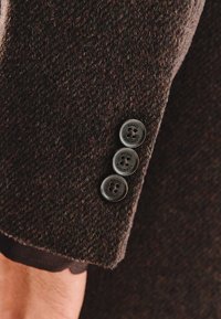 Teksturowana brązowa wełniana marynarka, z rękawem zdobionym trzema ciemnymi guzikami. Tkanina ma subtelne odcienie kolorystyczne i klasyczny krój.