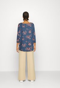 Person med flätat hår som vänder sig bort, klädd i en blå blommig långärmad topp, beige vida byxor och vita skor mot en vit bakgrund.