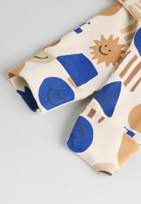 Tela de algodón con un fondo crema que presenta formas geométricas azules, soles sonrientes y varios patrones. Puños enrollados destacados.