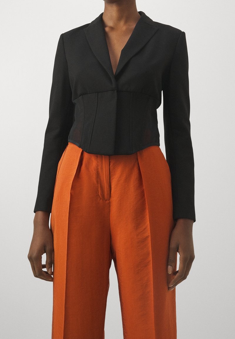 Cropped black blazer with a notched collar, featuring a fitted design and smooth texture, paired with high-waisted orange trousers.