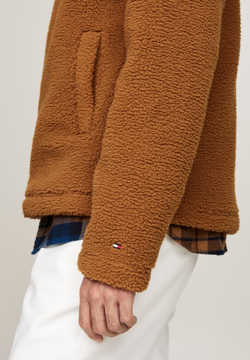 Giacca in pile marrone con superficie testurizzata, dotata di una tasca laterale e di una piccola patch con logo vicino al polsino. Indossata sopra una camicia a quadri.