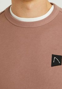 Sudadera rosa claro con cuello redondo, puños acanalados y un parche de logo negro con un contorno blanco. Tejido suave y texturizado.