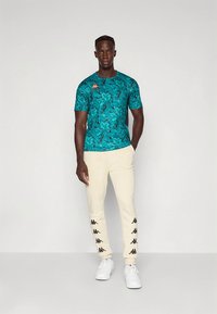 Tyrkis mønstret t-shirt med korte ærmer, kombineret med cremefarvede joggers med sorte logoaccent langs siderne. Hvide sneakers fuldender looket.