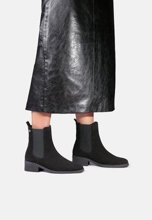 Jambes portant une jupe midi en cuir noir et des bottines Chelsea en daim noir avec des panneaux latéraux élastiques et des talons bas, debout sur un sol blanc.