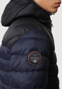 Veste matelassée bleu marine avec capuche, dotée d'un patch logo circulaire sur la manche gauche et d'une texture matelassée pour l'isolation.