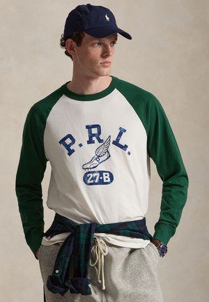 Man met een marineblauwe pet, wit shirt met lange mouwen en groene mouwen en grafische print, grijze joggingbroek, met een geruit overhemd gestrikt rond de middel.