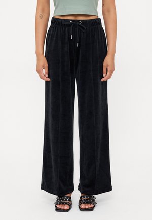 Persoon draagt zwarte velours broek met brede pijpen en een trekkoord in de taille, gecombineerd met open sandalen met meerdere riemen en studs.