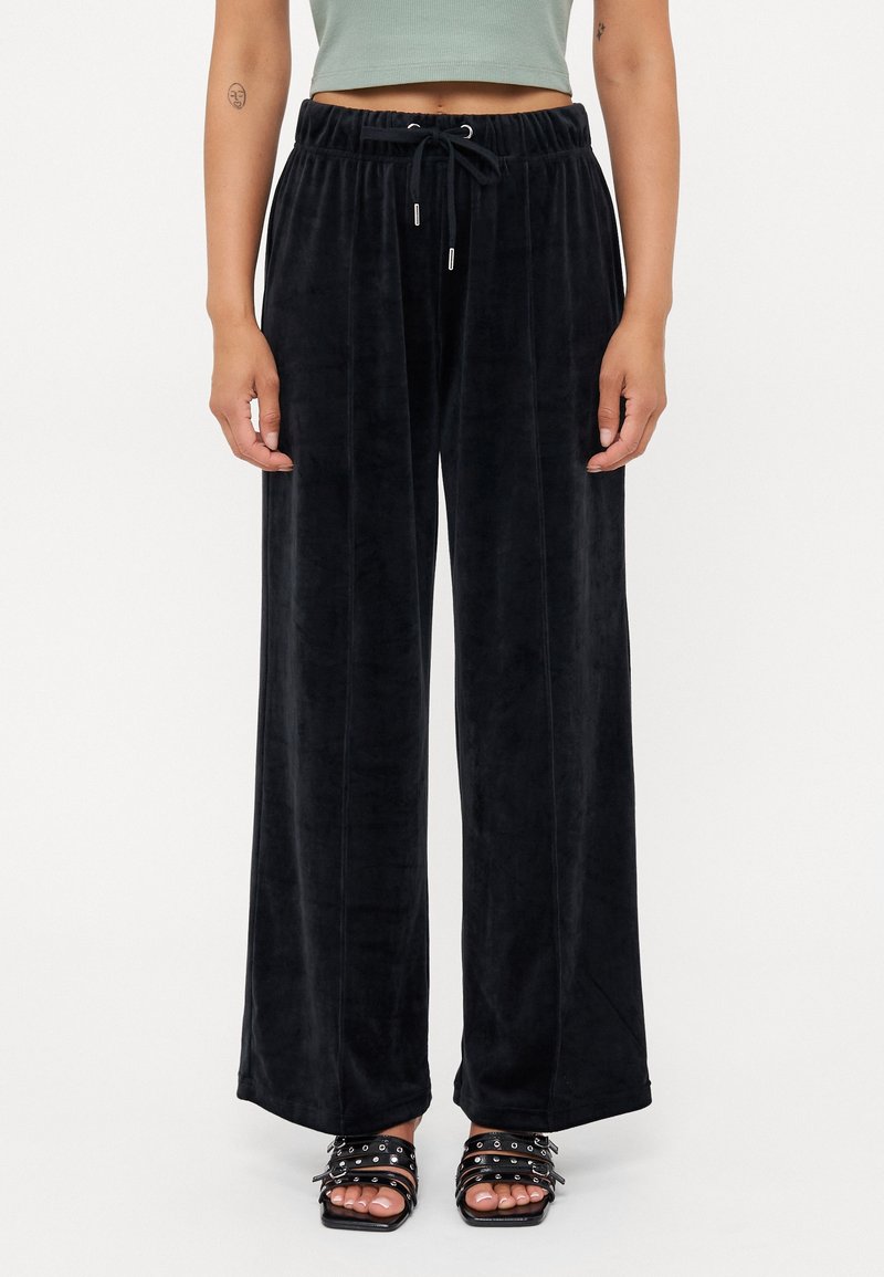Personne portant un pantalon large en velours noir avec une taille à cordon, associé à des sandales ouvertes avec plusieurs brides et clous.