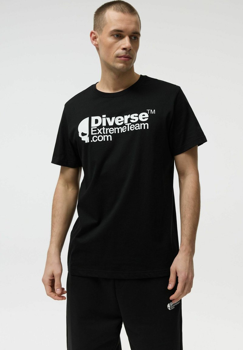 Młody mężczyzna w czarnej koszulce i spodniach z białym logo "Diverse ExtremeTeam.com", stojący na tle jasnego, gładkiego tła.