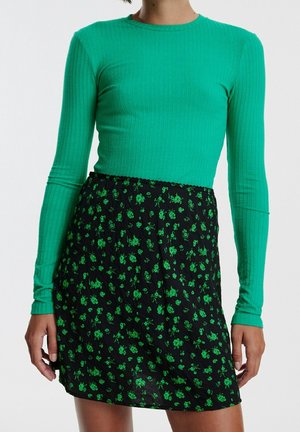 Groen geribbeld langslavend shirt gecombineerd met een zwarte rok met kleine groene bloemenpatronen. De outfit heeft een aansluitende pasvorm.