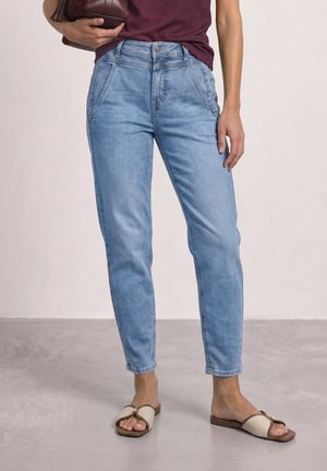 Femme portant un jean taille haute bleu clair, un top bordeaux, des sandales beiges et tenant un sac marron sur un fond clair uni.