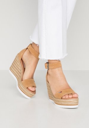 Fødder iført beige ruskind kilehælssandaler med anklerem og hvide bukser, der er cropped over anklen, mod en ensfarvet baggrund.