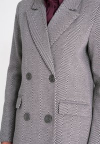 Dwurzędowa marynarka w kratę houndstooth w odcieniach szaro-czarnych z dużymi guzikami, klapami o wycięciu i bocznymi kieszeniami. Teksturowany wzór materiału.