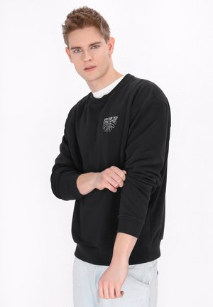 Hanorac negru cu mâneci lungi și manșete întărite, având un design grafic alb pe piept. Material moale cu o potrivire lejeră.
