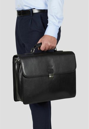 Mann i blå skjorte og mørke bukser holder en svart skinnveske med en sølvfarget lås i håndtaket mot en grå bakgrunn.